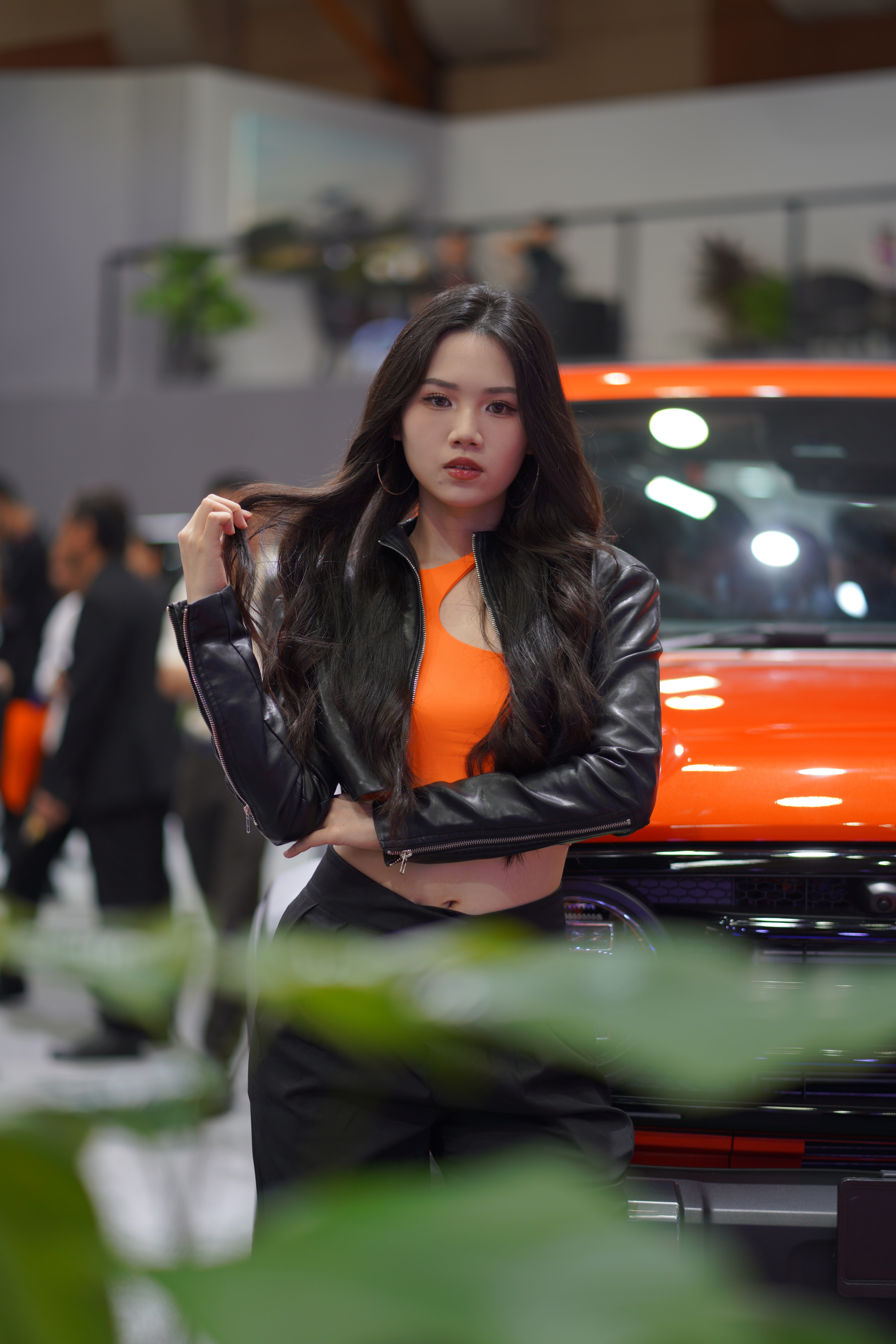 Model in a black jacket and orange top beside an orange car